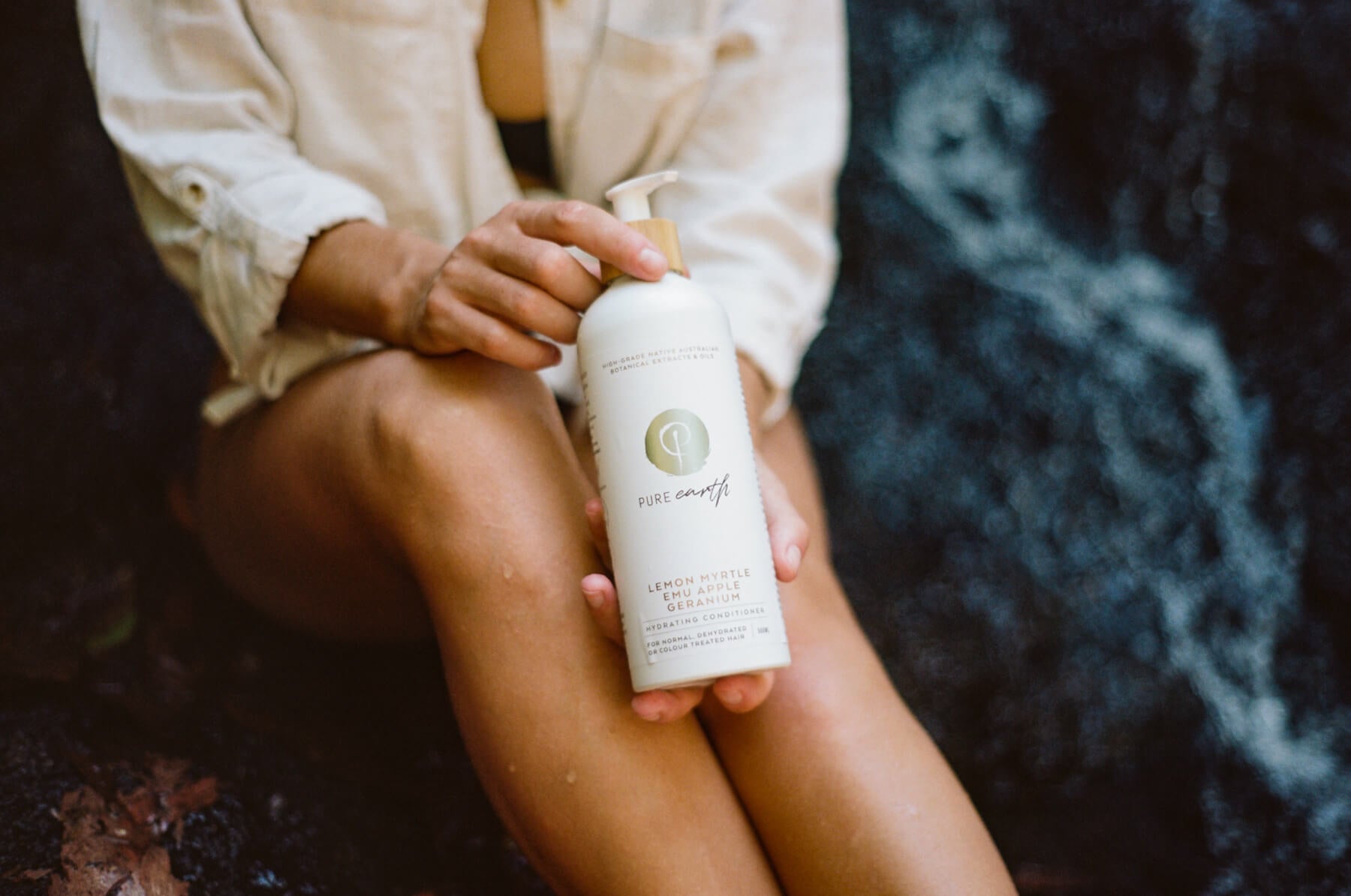 Woman holding white bottle with pump and label 'pure earth hydrating shampoo' in front of a blurred dark natural stone.