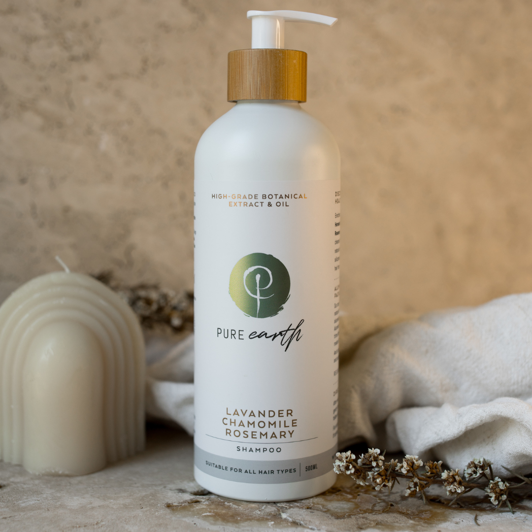 A bottle of Pure Earth Lavender Chamomile Botanical Shampoo with a pump dispenser, placed on a surface with a beige background. The bottle is white with a green and purple label and a wooden pump.