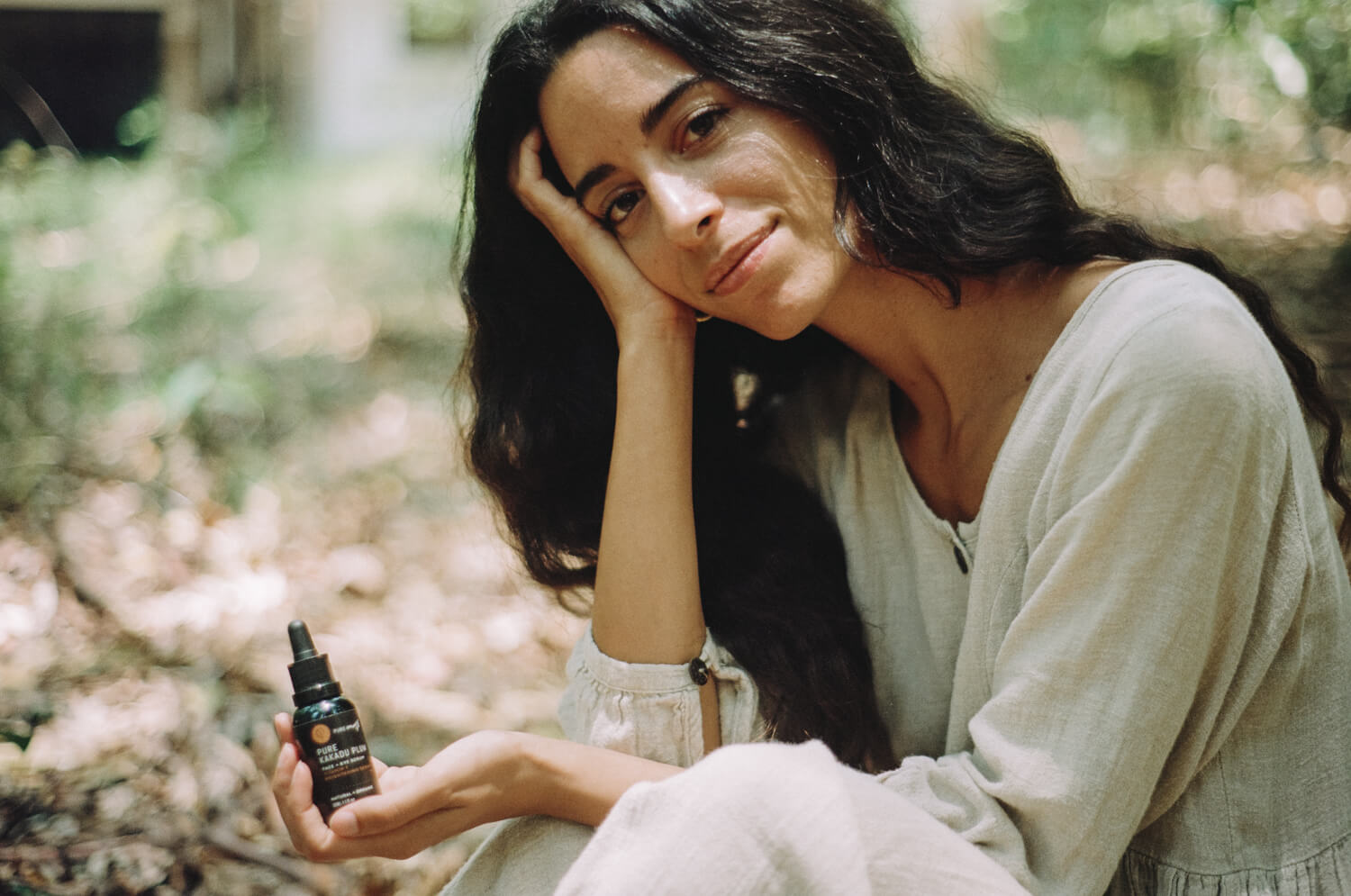 Woman in forest with linen clothes and long black hair holding a black droppper bottle with label 'Pure kakadu Plum Serum'