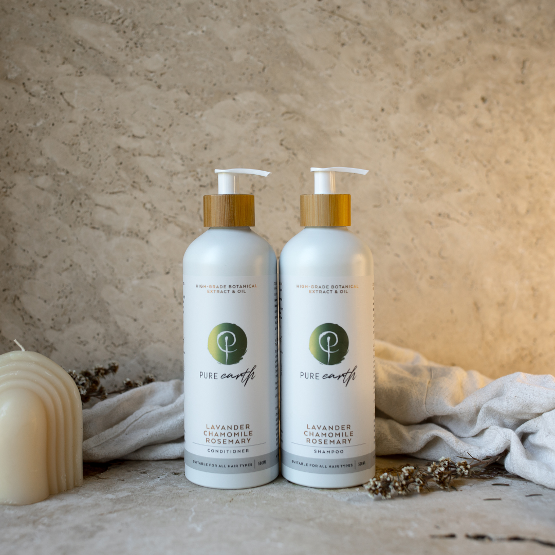 Two bottles of natural shampoo and conditioner with pump dispenser, placed on a surface with a neutral-toned background, accompanied by a candle and some dried flowers.