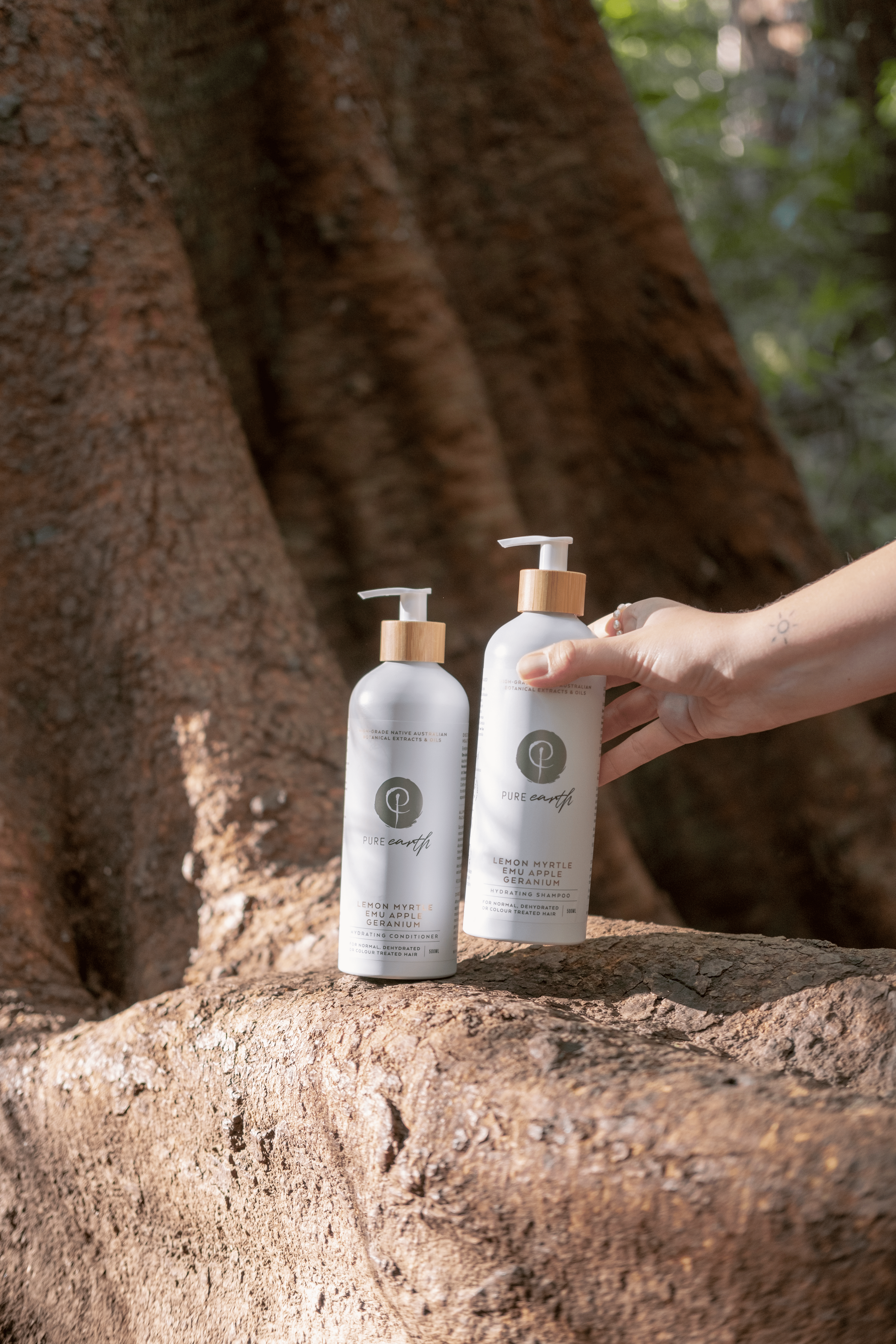 Two bottles of shampoo and conditioner  with label 'pure earth natural hydrating shampoo and conditioner' on a tree stump with a hand reaching for one bottle.