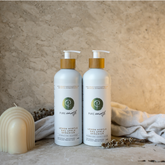 Two bottles of natural shampoo and conditioner with pump dispensers, displayed alongside a candle and some dried plants, against a textured beige background.