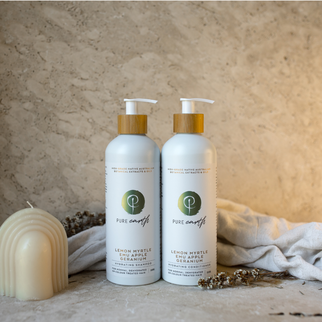 Two bottles of natural shampoo and conditioner with pump dispensers, displayed alongside a candle and some dried plants, against a textured beige background.