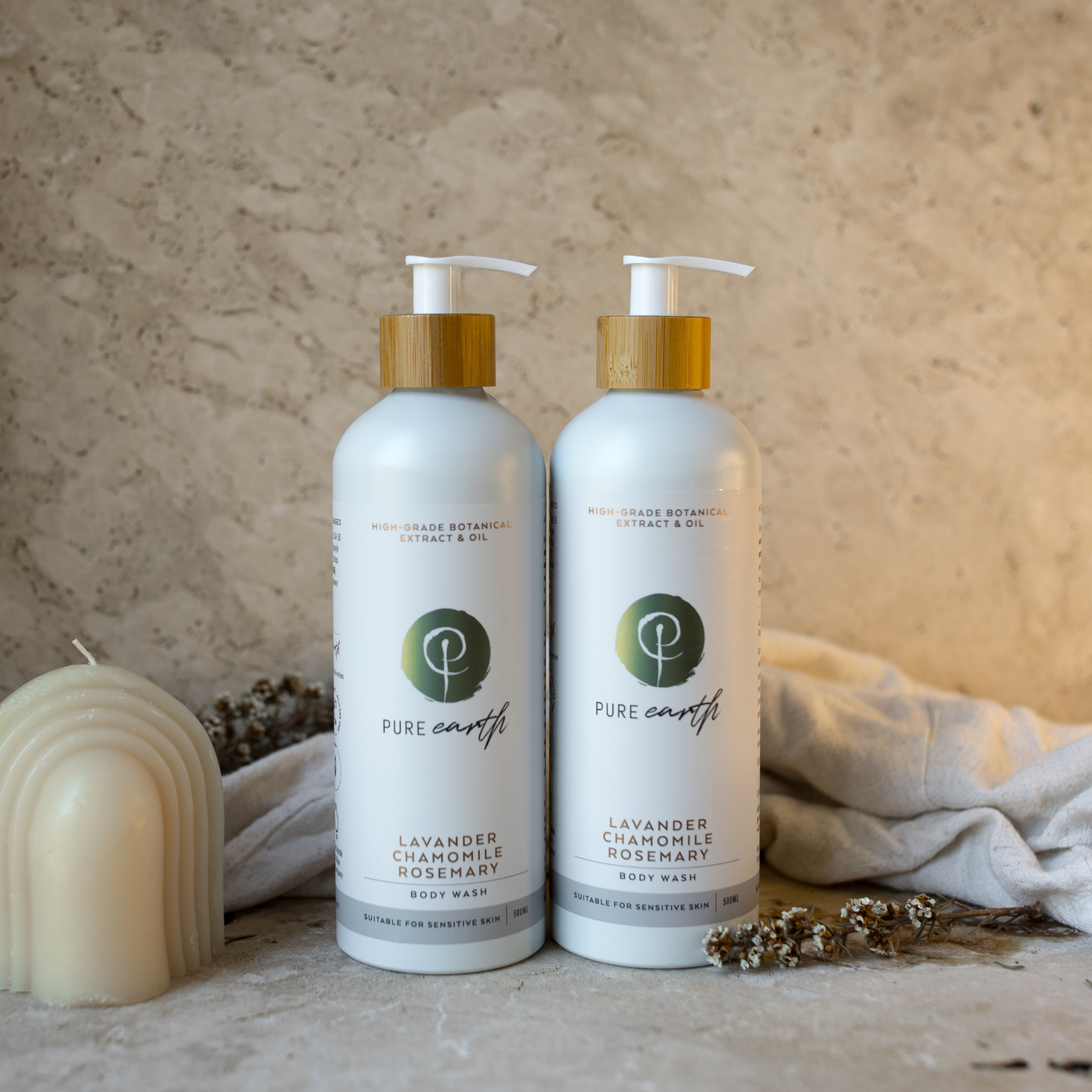 Two bottles of Pure Earth body wash on a wooden surface with a candle and towel in the background.